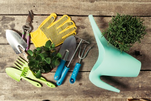 The image displays a collection of gardening tools and accessories arranged on a weathered wooden surface. Included are yellow rubber gloves with a textured grip, a small hand trowel with a metallic blade, a pair of pruners with purple handles, a pitchfork with a metal head and blue handle, and a green-handled tool. There is also a light turquoise plastic watering can with a long spout, and a potted plant with dense green foliage, likely a herb such as rosemary. The wooden surface provides a rustic background, highlighting the outdoor gardening theme. This setup suggests preparations for gardening activities typical of a well-maintained outdoor space in Hampstead or similar London suburbs. The arrangement and choice of tools are indicative of general gardening, hedge trimming, and lawn care tasks that a professional gardening service like Hedge Trimming Hampstead might undertake for local clients, emphasizing attention to detail and quality outdoor maintenance in residential or communal gardens during suitable weather conditions.