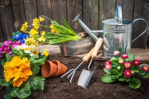 An outdoor garden scene featuring freshly turned dark soil in the foreground, ready for planting, with various gardening tools such as a small garden fork, trowel, and watering can resting on the soil. To the left, vibrant flowering plants including yellow primroses, purple pansies, and white and yellow daffodils are in full bloom, adding bright colour contrasts to the scene. Behind these, a set of empty terracotta pots are stacked on the ground, indicating ongoing planting activities. The background consists of a weathered wooden fence, providing a natural boundary for the garden space. The overall environment suggests a well-maintained garden in a residential area near Hampstead, with natural sunlight illuminating the scene, creating a welcoming and productive outdoor setting suitable for gardening and landscaping services by Hedge Trimming Hampstead.