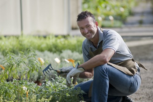 A man with short dark hair and a friendly smile is kneeling in a well-maintained garden, using a garden fork to tend to lush green plants and shrubs. The garden features a neatly trimmed lawn in the foreground, bordered by flower beds with a variety of foliage. In the background, there are small trees and a wooden fence, indicating a private outdoor space typical of suburban Hampstead. The man is dressed in casual clothing with a light grey shirt, blue jeans, and a gardening apron, suggesting he is engaged in gardening or landscaping work. The environment is bright and airy, with natural daylight illuminating the scene and highlighting the vibrant green tones of the plants. This image reflects professional gardening care in a typical London garden setting, aligning with services offered by Hedge Trimming Hampstead for maintaining and enhancing outdoor landscaped areas, including hedge trimming, lawn care, and general garden maintenance in the Hampstead area through the post code NW3.