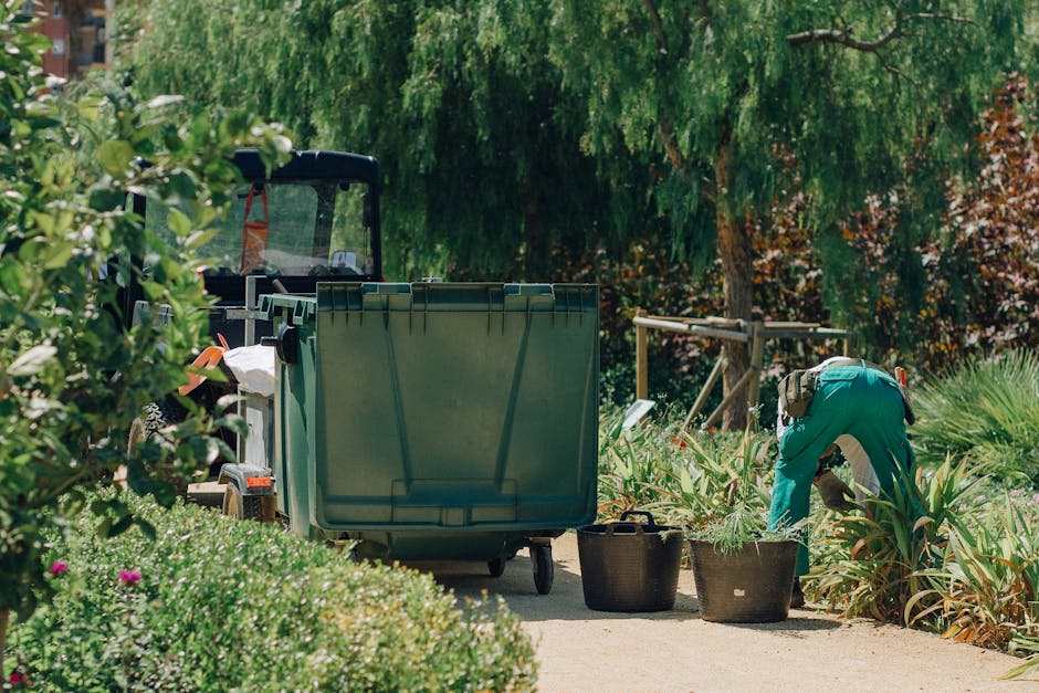 A gardener dressed in teal overalls is crouched down, trimming plants in a lush garden area with a pair of pruning shears. To the left, a large green waste bin on wheels is positioned on a concrete pa