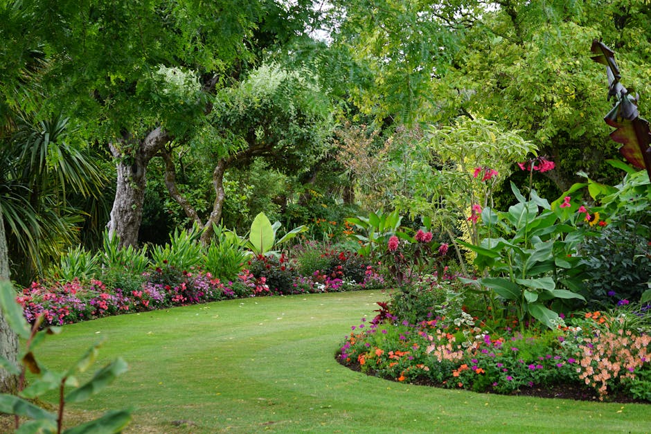 A gardener dressed in teal overalls is crouched down, trimming plants in a lush garden area with a pair of pruning shears. To the left, a large green waste bin on wheels is positioned on a concrete pa