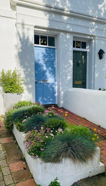 A neatly maintained front garden featuring a paved stone patio with a central pathway leading to a decorative wrought iron bench positioned against a white exterior wall of a house. The garden is bord